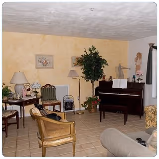 A cozy living room with a piano against a yellow textured wall, a tall green plant beside it, and various chairs including a wicker chair and cushioned seats arranged around the room. There are framed pictures on the wall, a floor lamp, and a small table with a lamp and decorative items.