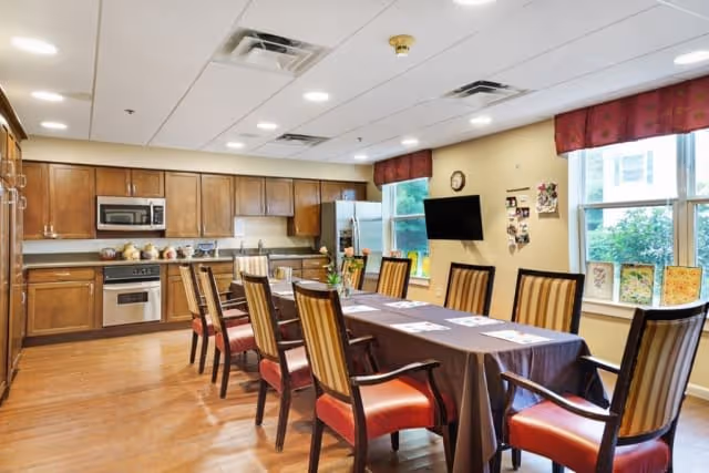 A spacious kitchen and dining area with wooden cabinets, stainless steel appliances including a microwave and oven, and a long dining table covered with a dark tablecloth surrounded by wooden chairs with red cushions. The room has large windows with red valances, a wall-mounted TV, and various decorations on the walls and windowsills.
