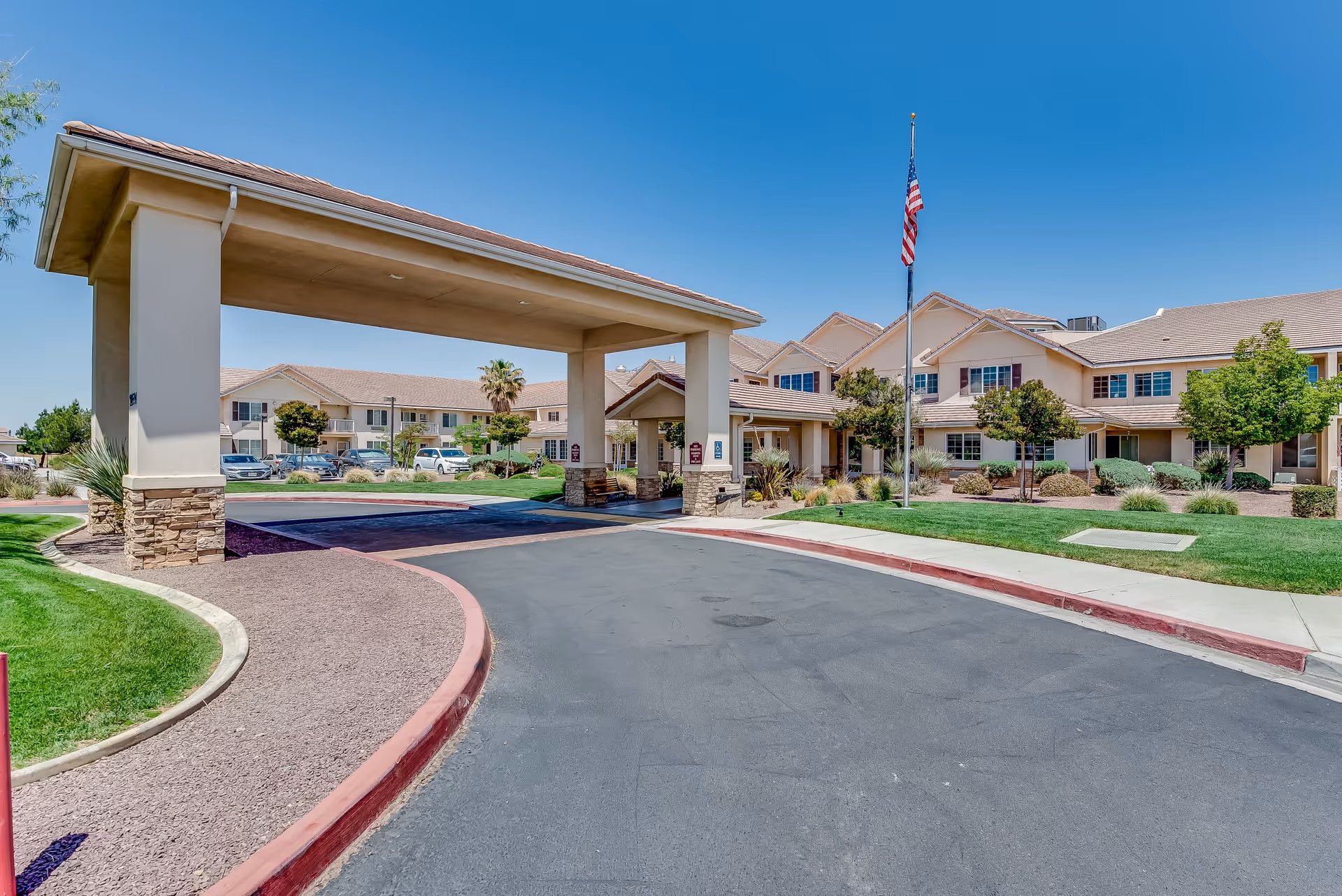 Exterior view of Solstice Senior Living at Apple Valley showing the main entrance with a covered drop-off area, a flagpole with an American flag, landscaped greenery, and a clear blue sky.