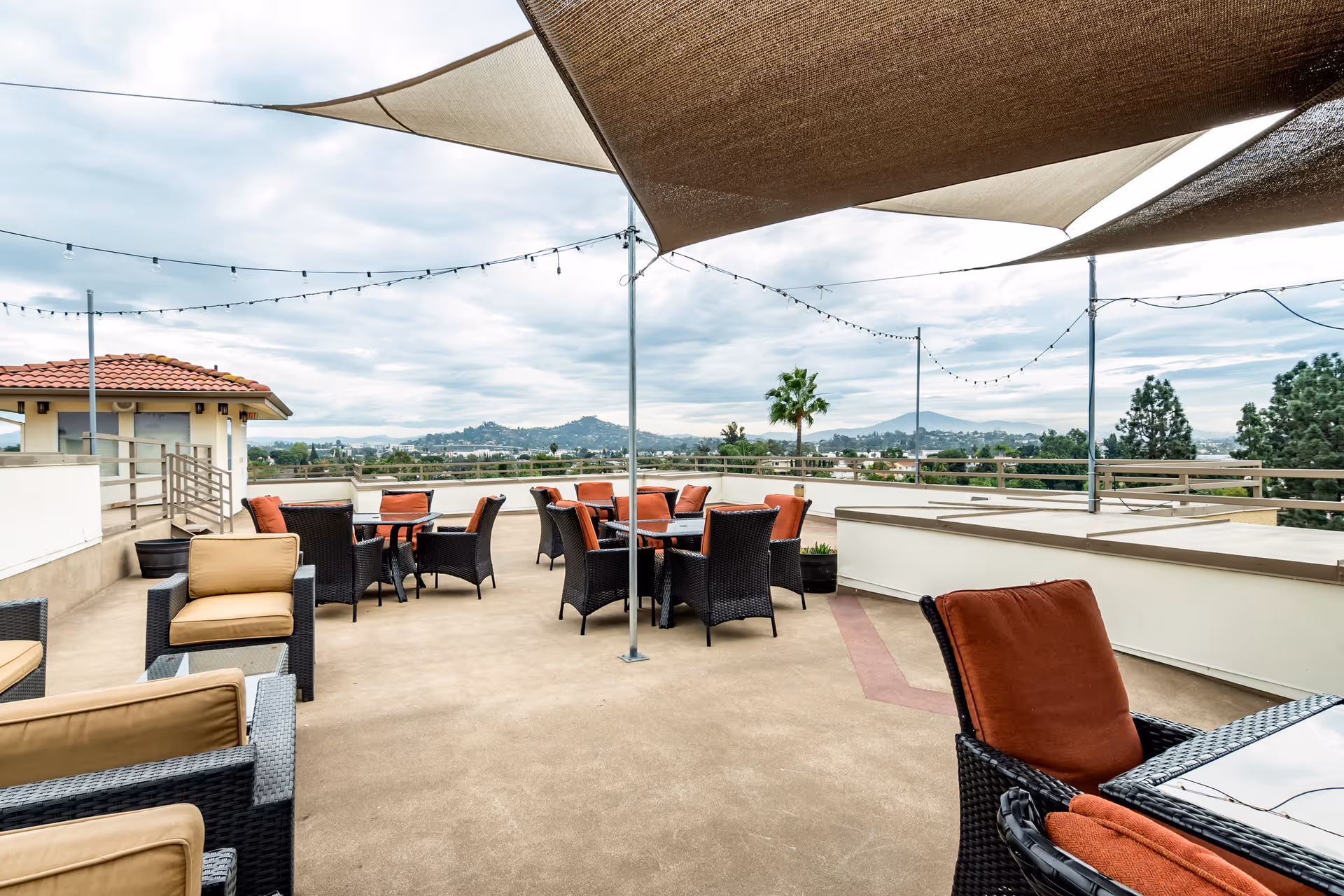 Outdoor patio area with multiple wicker chairs and tables, some with orange cushions and others with beige cushions. Overhead, there are triangular shade sails and string lights. The patio overlooks a scenic view of distant hills and trees under a cloudy sky.