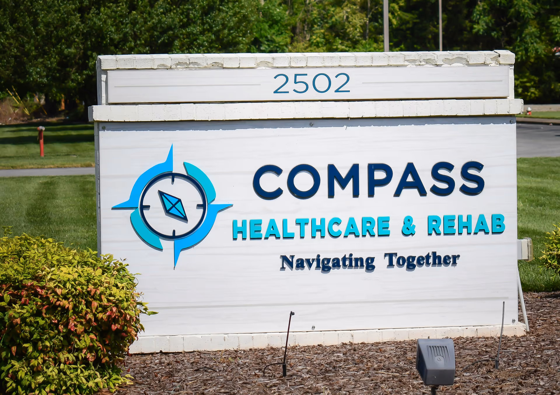 Outdoor sign for Compass Healthcare & Rehab with the address number 2502 displayed above. The sign includes a compass logo and the tagline 'Navigating Together'. The sign is surrounded by greenery and landscaping.
