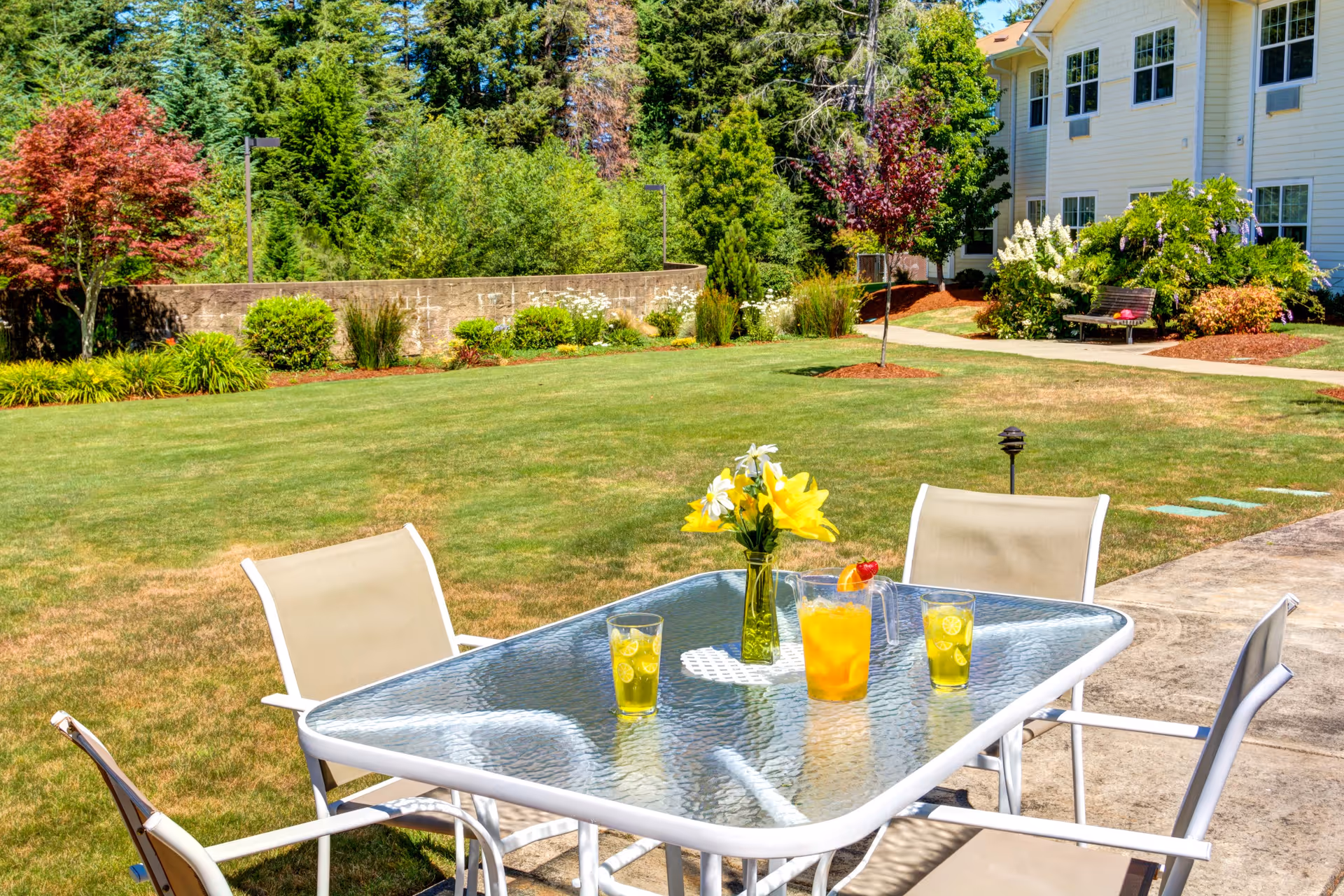 Outdoor patio area with a glass table and four beige chairs. On the table, there is a vase with yellow and white flowers, a pitcher of orange drink with a strawberry garnish, and three glasses filled with a yellow beverage. The patio overlooks a well-maintained lawn with various shrubs, trees, and a bench near a white building in the background.