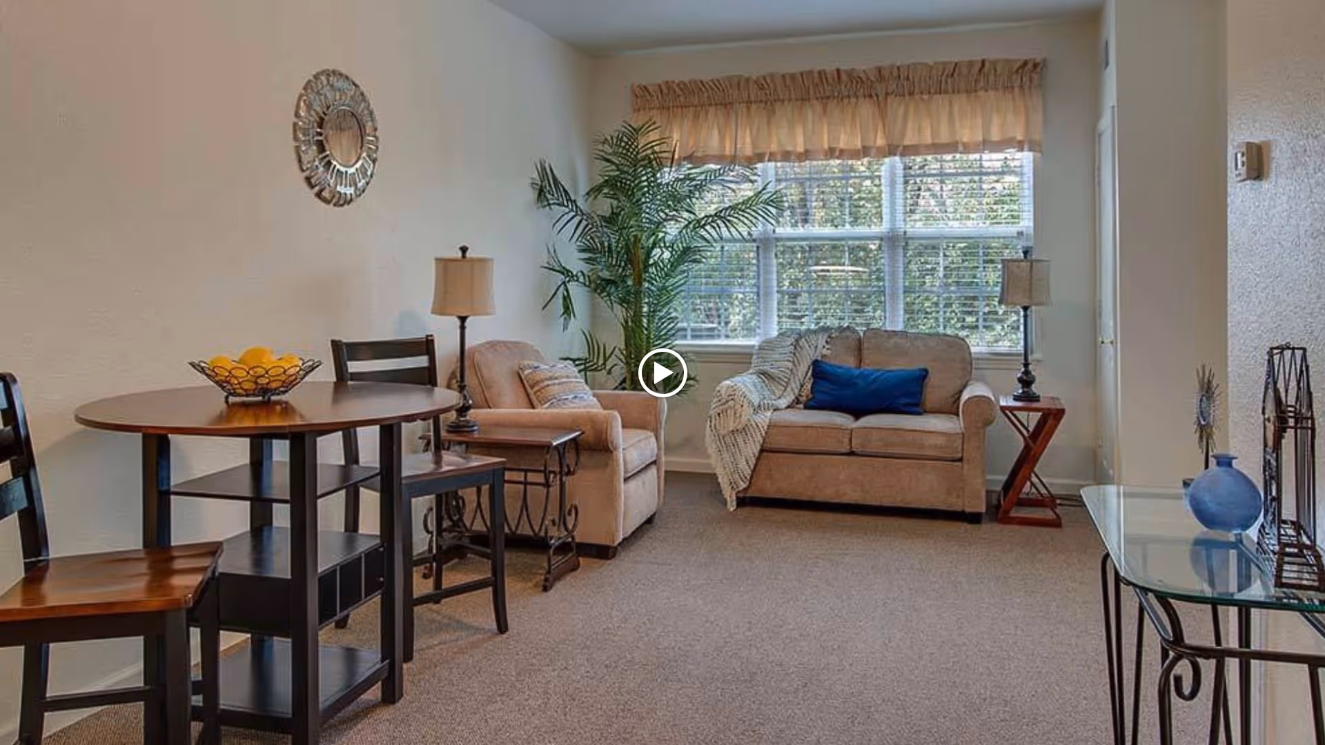 A bright living room with a loveseat and armchair by a large window, a small dining table with chairs, lamps, and decorative plants and accessories.