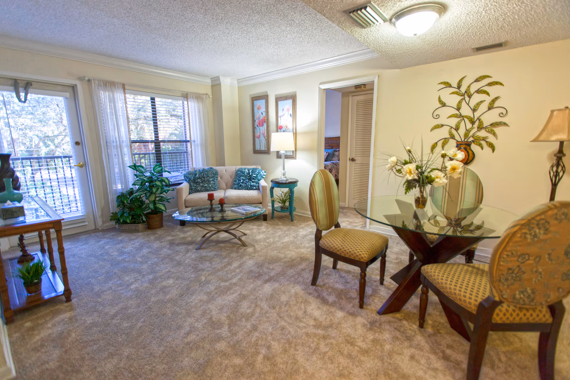 A cozy living room with beige carpet, a small white loveseat with blue pillows, a glass coffee table, and a round glass dining table with three upholstered chairs. There are two framed floral paintings on the wall, a floor lamp, a side table with a lamp, and decorative plants near a window and glass door leading outside.