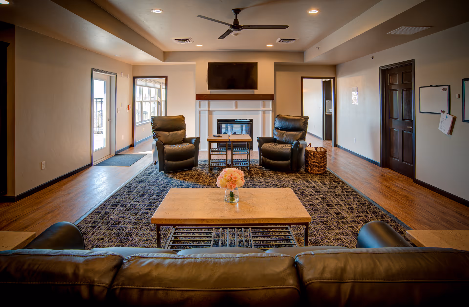 A cozy living room area with a dark leather sofa in the foreground, a rectangular coffee table with a small vase of flowers, two black recliner chairs facing a fireplace with a mounted flat-screen TV above it. The room has wooden flooring, a patterned area rug, and neutral-colored walls with doorways leading to other rooms and a glass door to the outside.
