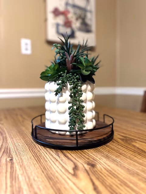 A close-up of a white textured ceramic pot with various green succulent plants inside, placed on a round black metal tray on a wooden table. The background shows a beige wall with white trim and a blurred framed picture.