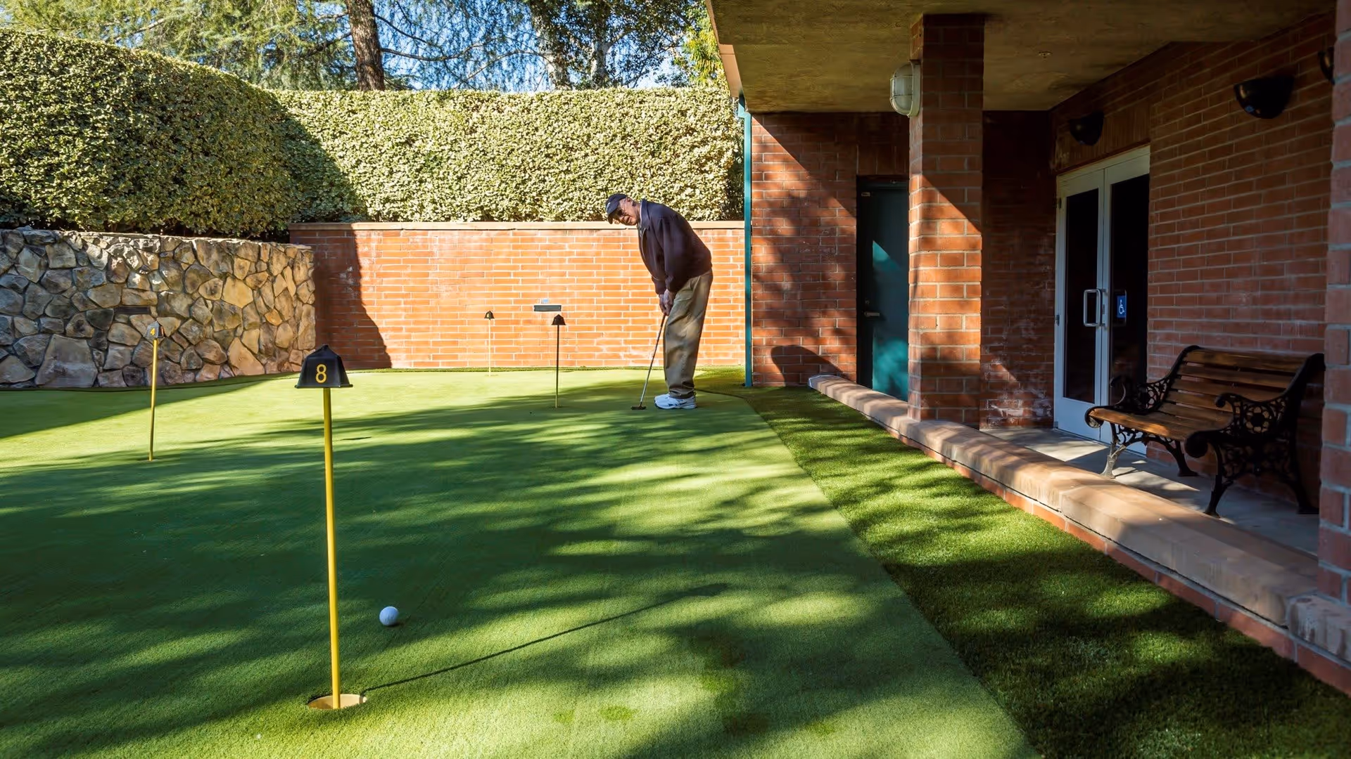 A person putting on a small outdoor putting green next to a brick building and bench.
