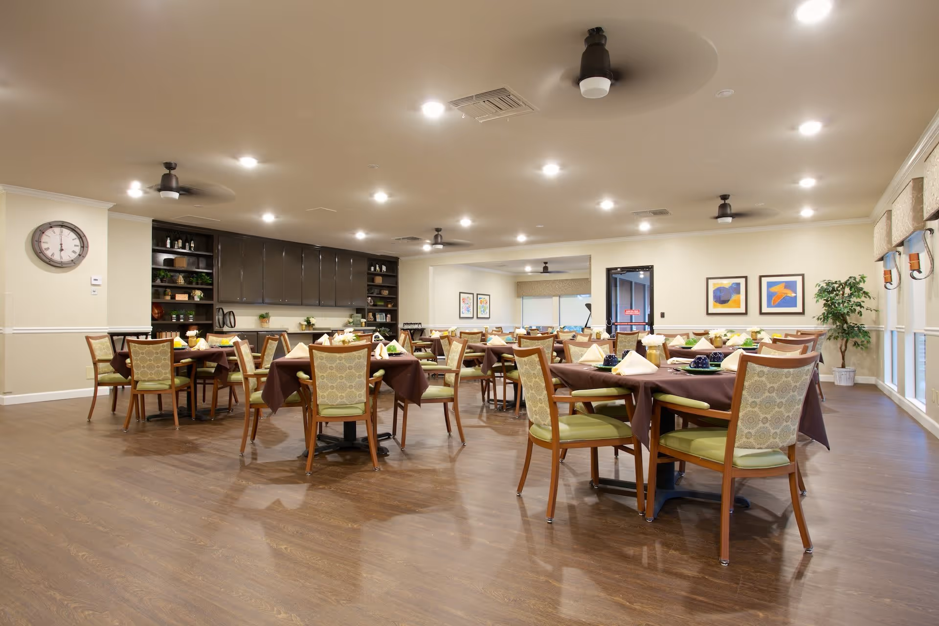 A spacious dining room with multiple round tables covered with brown tablecloths and set with napkins and cups. The room has wooden flooring, beige walls, ceiling fans, recessed lighting, and large windows with valances. There are decorative plants, framed artwork on the walls, and built-in cabinets with shelves along one side.