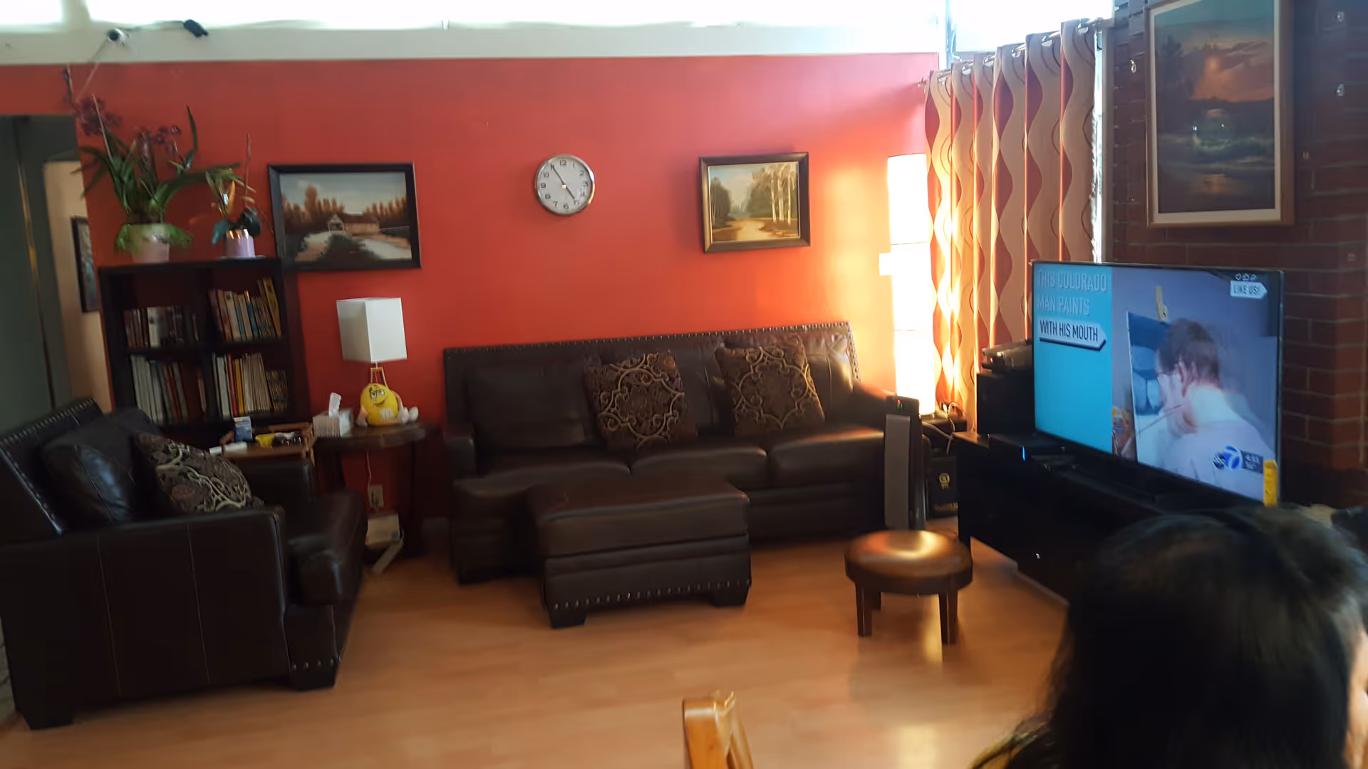 Living room with dark leather sofas and ottoman, a TV on a stand, a red accent wall with paintings and a clock, and a bookshelf with plants.