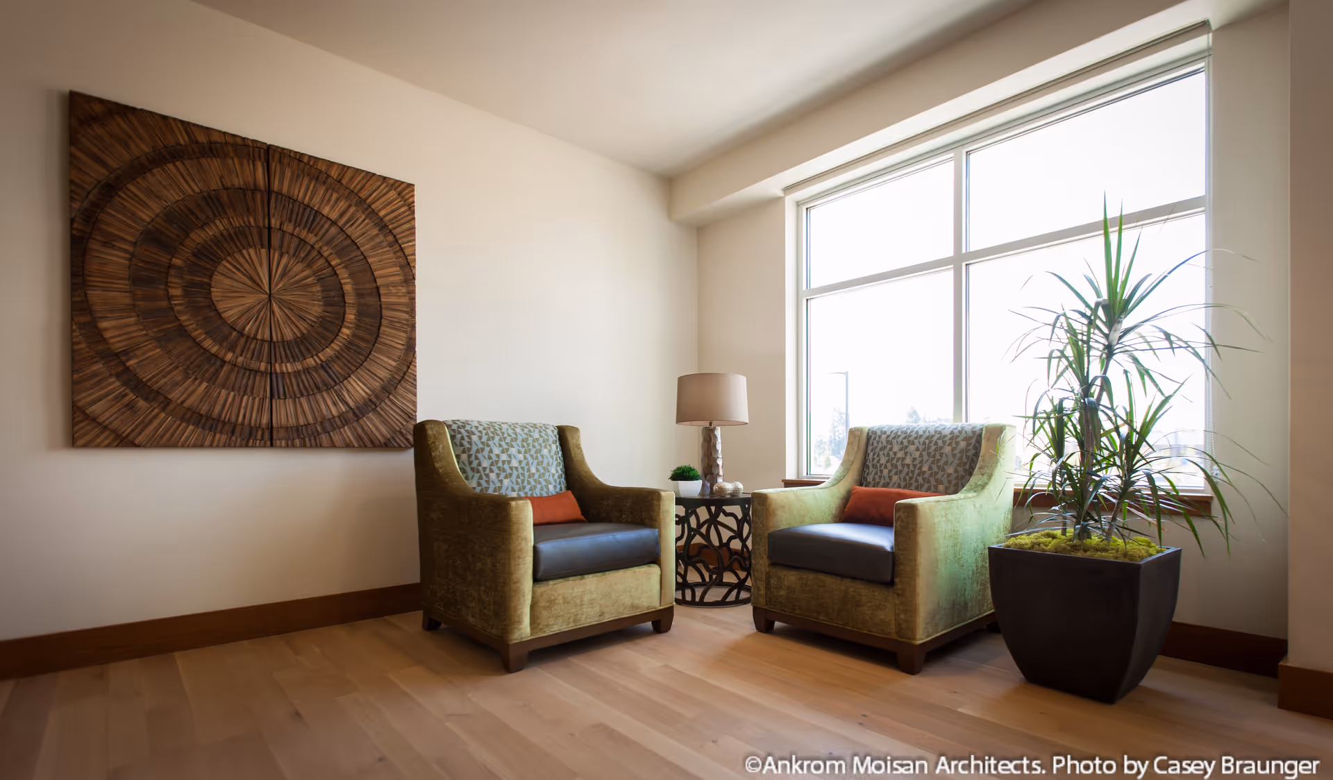 A cozy seating area with two green upholstered armchairs featuring patterned backs and orange accent pillows, positioned around a small round table with a lamp and a small plant. A large window lets in natural light, and a tall potted plant is placed beside the chairs. A decorative wooden wall art piece hangs on the adjacent wall.