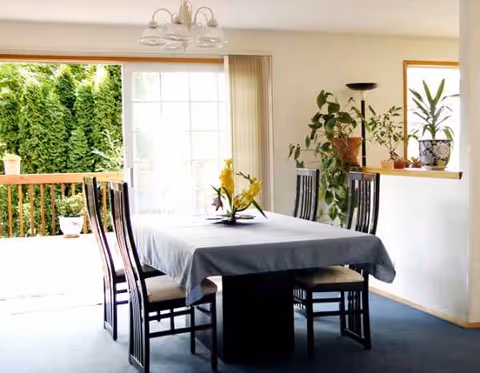 Indoor dining room in a senior living facility with a table, chairs, plants, and windows.