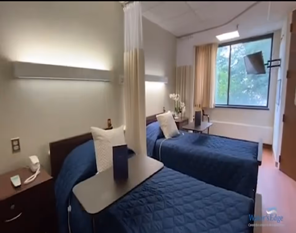 A bright rehabilitation patient room with two single beds covered in navy quilts, separated by a privacy curtain, with over-bed tables, nightstands, and a window.