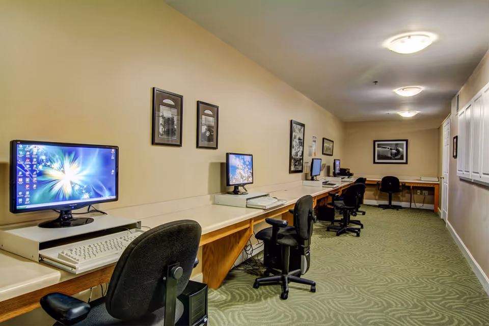 A long interior computer/business center with a row of desks, desktop computers, and office chairs along a carpeted hallway.