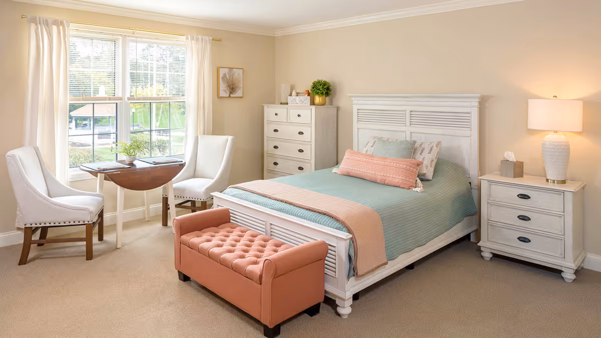 A cozy bedroom with a white wooden bed featuring a teal bedspread and pink throw blanket, a matching white dresser and nightstand with a lamp, a pink tufted bench at the foot of the bed, two white upholstered chairs, and a small wooden table near a window with white curtains.