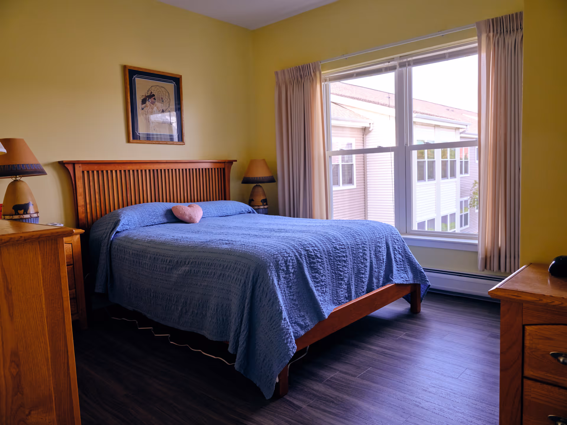 A bedroom with a wooden bed frame and a blue quilted bedspread. There is a small heart-shaped pillow on the bed. The room has yellow walls, a large window with beige curtains, and wooden furniture including nightstands with matching lamps on either side of the bed. A framed picture hangs on the wall above the headboard.