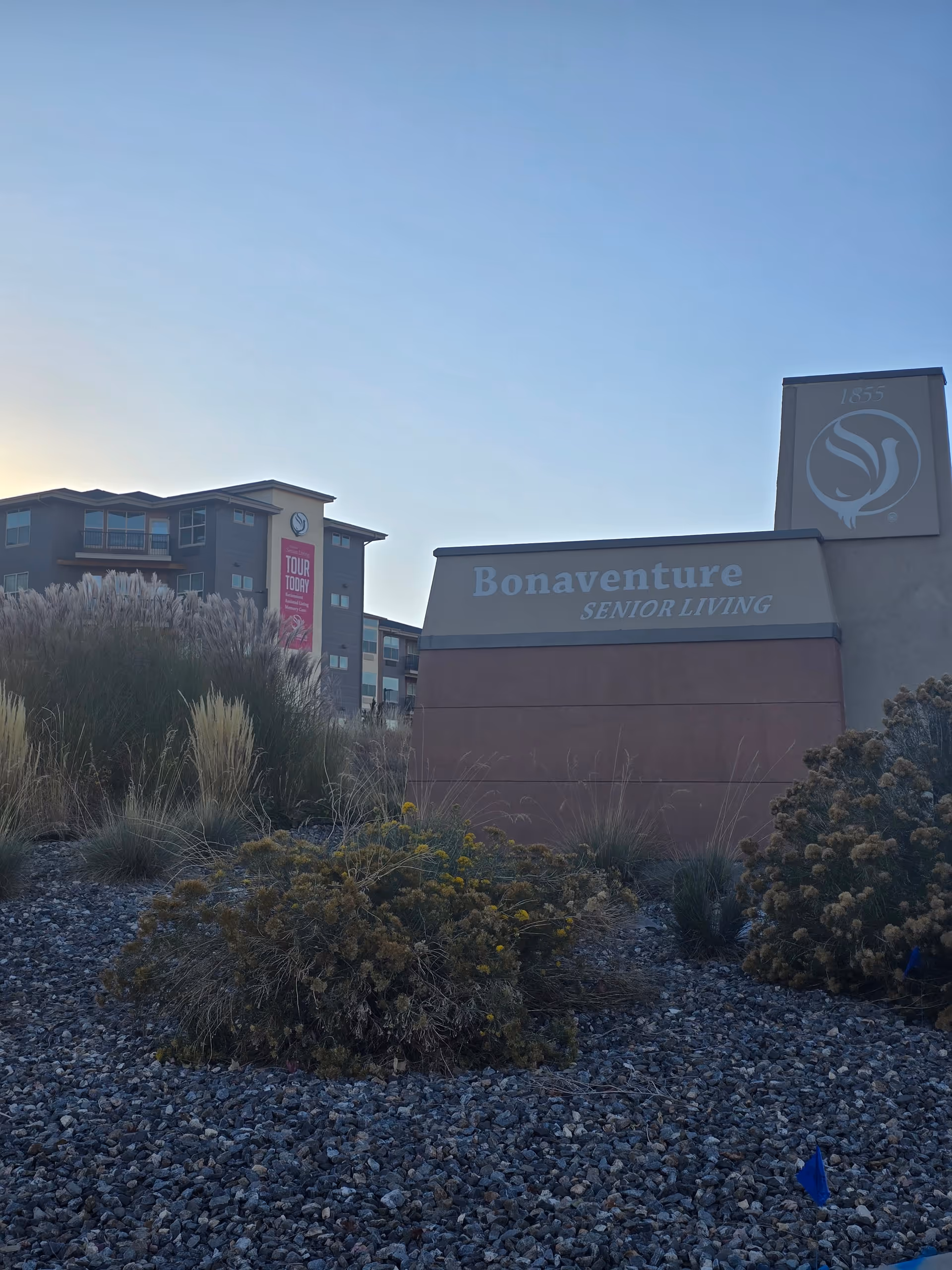 Exterior view of Bonaventure Senior Living facility with a large sign displaying the name. The building is visible in the background with some ornamental grasses and shrubs in the foreground under a clear sky.