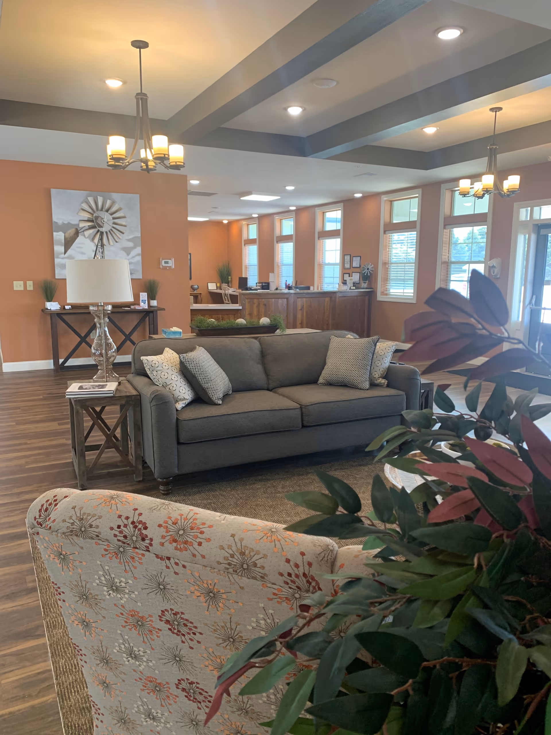 A cozy living room area in a retirement facility featuring a gray sofa with patterned cushions, a floral upholstered armchair, a side table with a lamp, and a console table with decorative items. The room has warm orange walls, wooden flooring, multiple windows letting in natural light, and ceiling lights with chandeliers. There is a reception desk visible in the background.