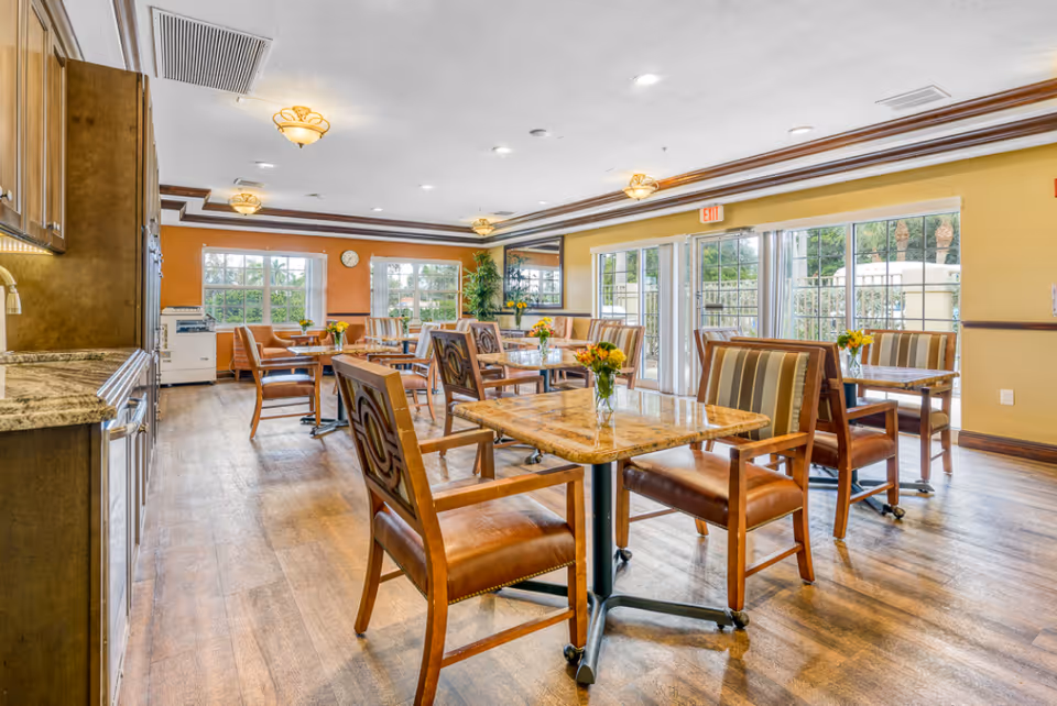 A bright and spacious dining room in a senior living facility with wooden tables and chairs arranged neatly. Each table has a small vase with yellow flowers. Large windows and glass doors allow natural light to fill the room, and the walls are painted in warm tones of yellow and orange. The floor is wooden, and ceiling lights provide additional illumination.