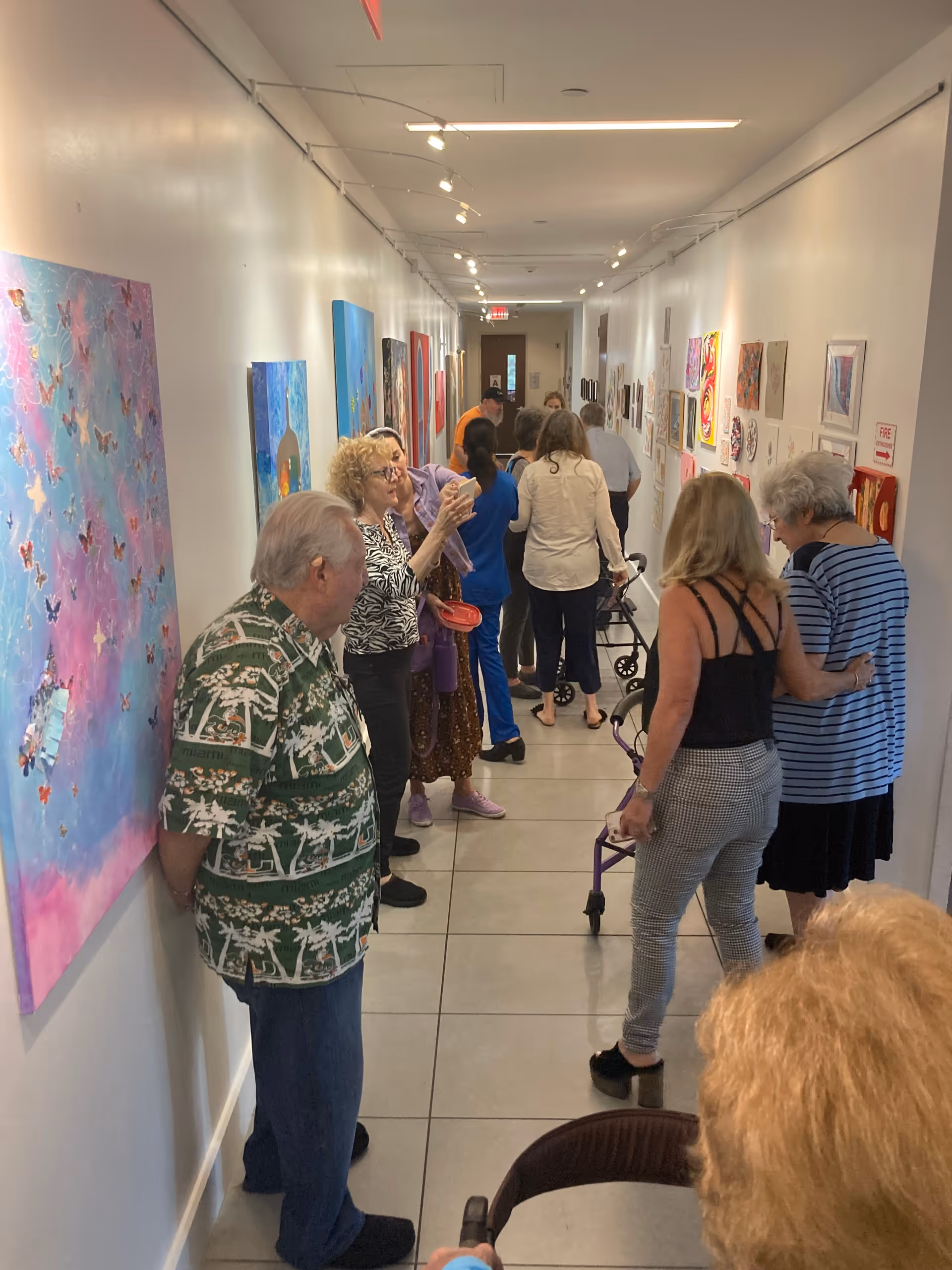 A group of elderly people and caregivers gathered in a brightly lit hallway with white walls adorned with colorful artwork. Some individuals are using walkers, and others are standing and conversing while observing the art pieces.