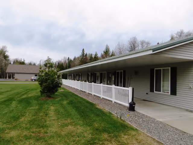 Exterior view of a single-story building with a long covered porch and white railing, surrounded by green grass and trees under a cloudy sky.