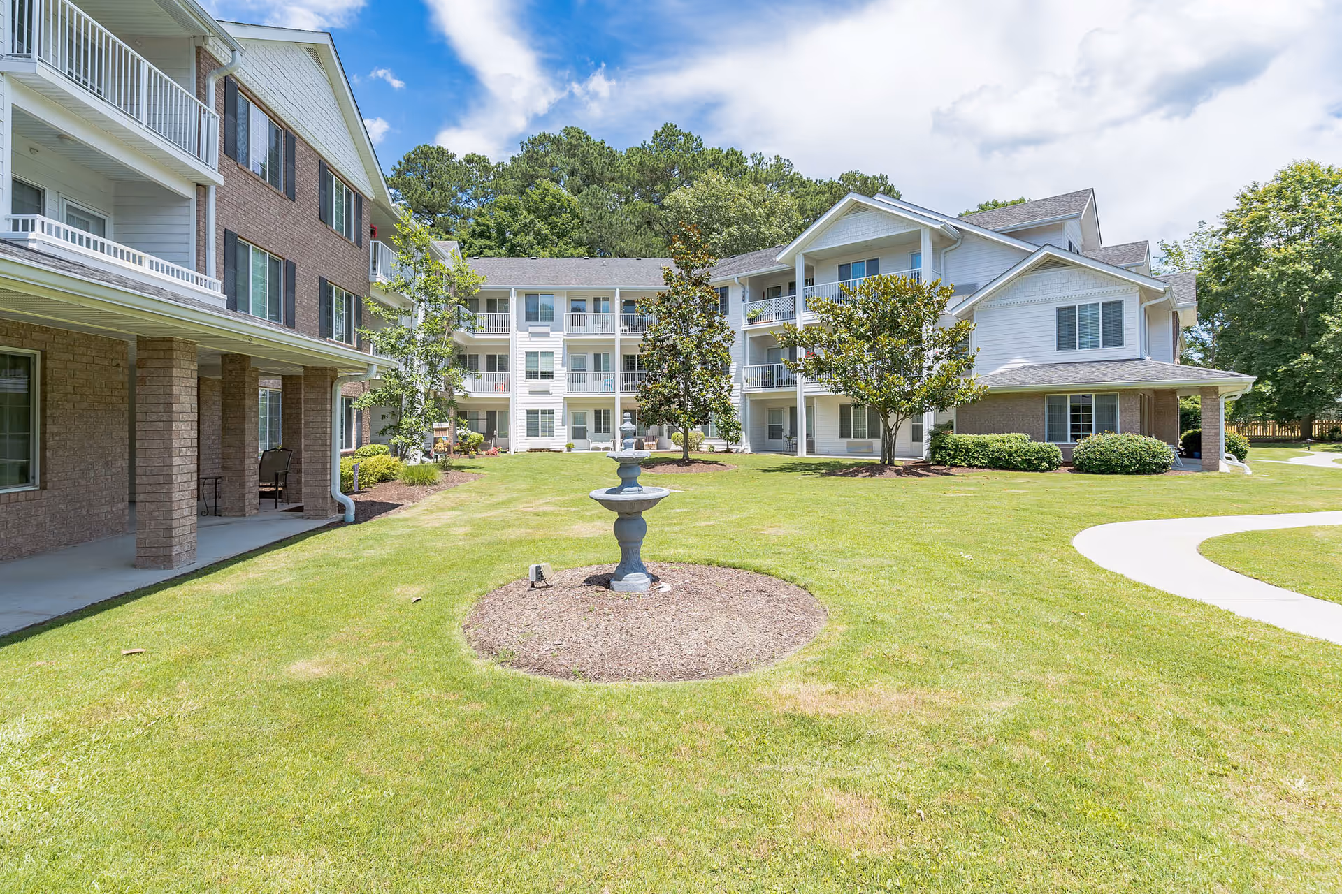 Outdoor view of a senior living facility with a well-maintained lawn, a small fountain in the center, several trees, and a three-story building with balconies and windows under a partly cloudy sky.