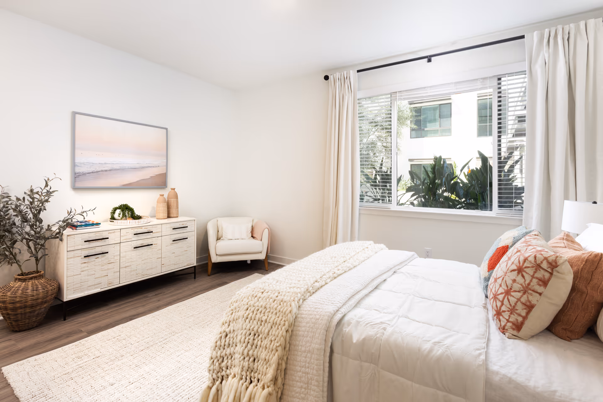 A bright and cozy bedroom featuring a neatly made bed with white bedding and decorative pillows in shades of orange and beige. A large window with white curtains lets in natural light and shows greenery outside. There is a white armchair in the corner and a white dresser with black handles against the wall, decorated with vases, books, and a small plant. A framed picture of a beach scene hangs above the dresser. The room has wooden flooring and a light-colored area rug.