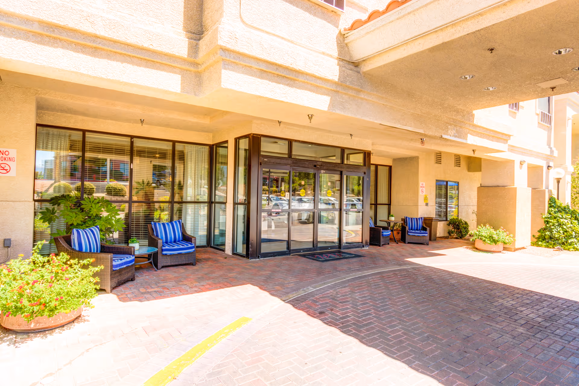 Entrance area of a building with glass double doors, outdoor seating with wicker chairs and blue striped cushions, potted plants, and a brick-paved driveway under a covered overhang.