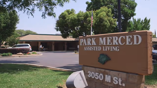 Front entrance sign reading "PARK MERCED ASSISTED LIVING" with the building, driveway, and trees in the background.