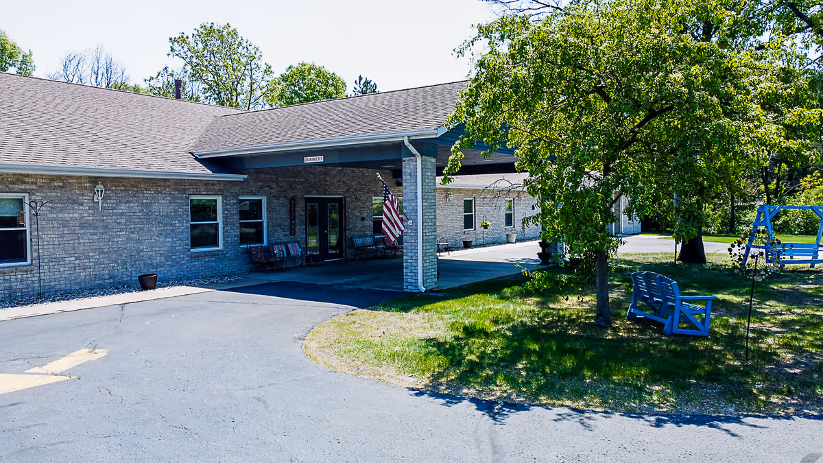 Exterior view of a single-story brick building with a covered entrance. There are benches and an American flag near the entrance. The surrounding area includes green grass, trees, and outdoor seating including a blue bench and a blue swing.