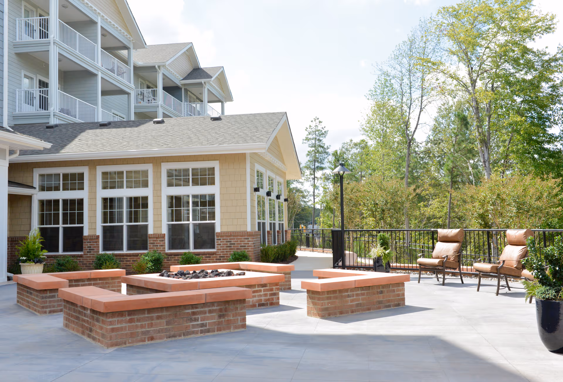 Outdoor patio area at a retirement living facility with brick fire pit surrounded by built-in benches with terracotta tops, cushioned chairs, potted plants, and a backdrop of trees and a multi-story building with balconies and large windows.