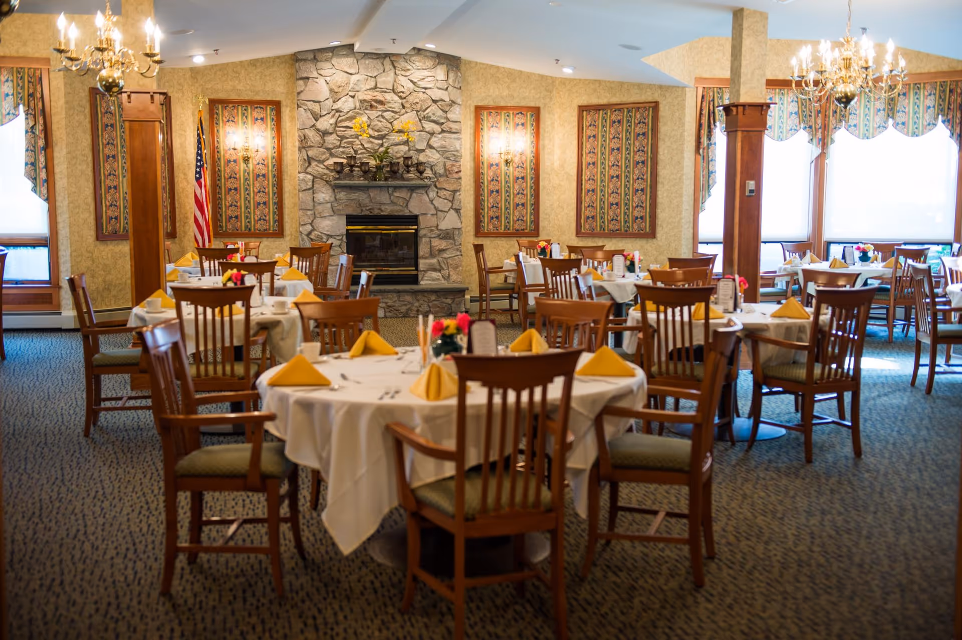 Elegant dining room with round tables set with white tablecloths, yellow napkins, wooden chairs, and a stone fireplace.