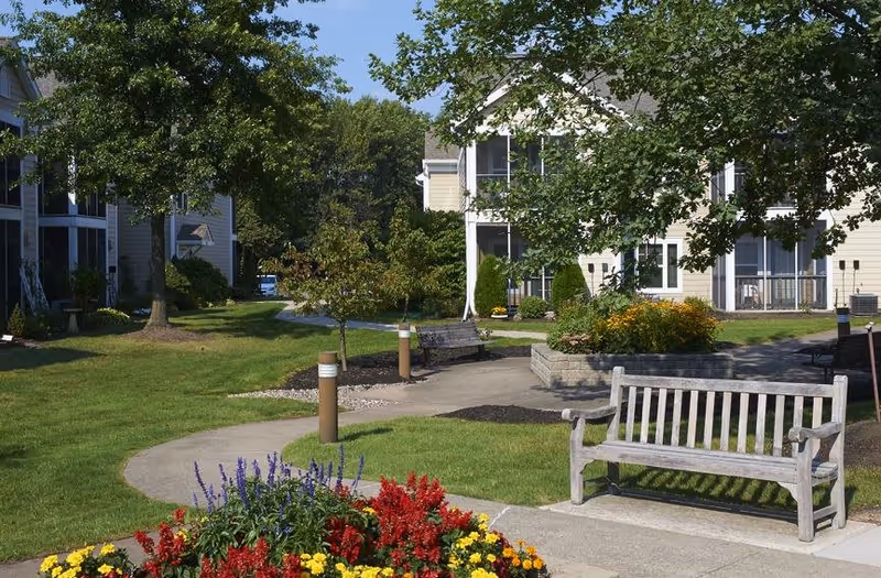 Outdoor garden area with a curved concrete walkway, wooden benches, colorful flower beds, and trees, surrounded by beige residential buildings under a clear blue sky.