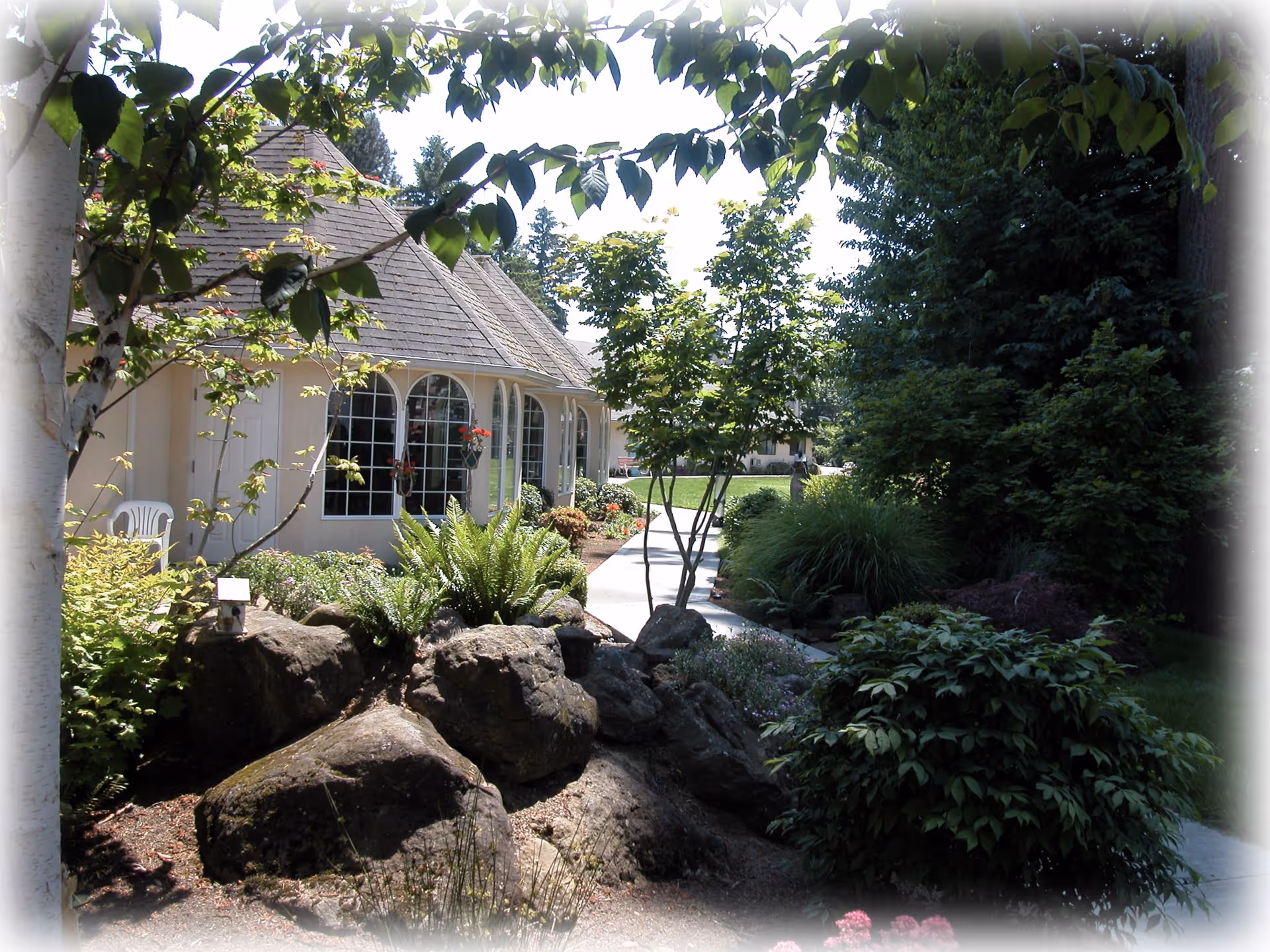 A landscaped outdoor area of a senior living facility featuring a walkway surrounded by rocks, bushes, trees, and various plants. A building with large arched windows and a shingled roof is visible on the left side, with a white plastic chair near the wall. The scene is bright and sunny with greenery providing a peaceful garden-like environment.