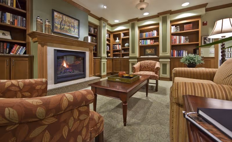 Cozy living room area with a lit fireplace surrounded by wooden bookshelves filled with books and decorative items. The room features comfortable upholstered armchairs and a sofa around a wooden coffee table with a tray and decorative objects. Warm lighting and a framed painting above the fireplace add to the inviting atmosphere.