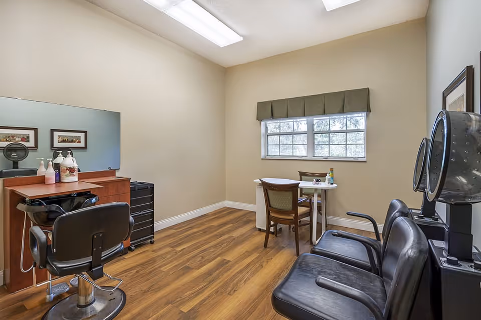 Interior view of a small salon room with a hair styling chair in front of a mirror, a wooden cabinet with hair care products, a black rolling cart, two black salon dryer chairs, and a small table with two chairs near a window with a green valance.
