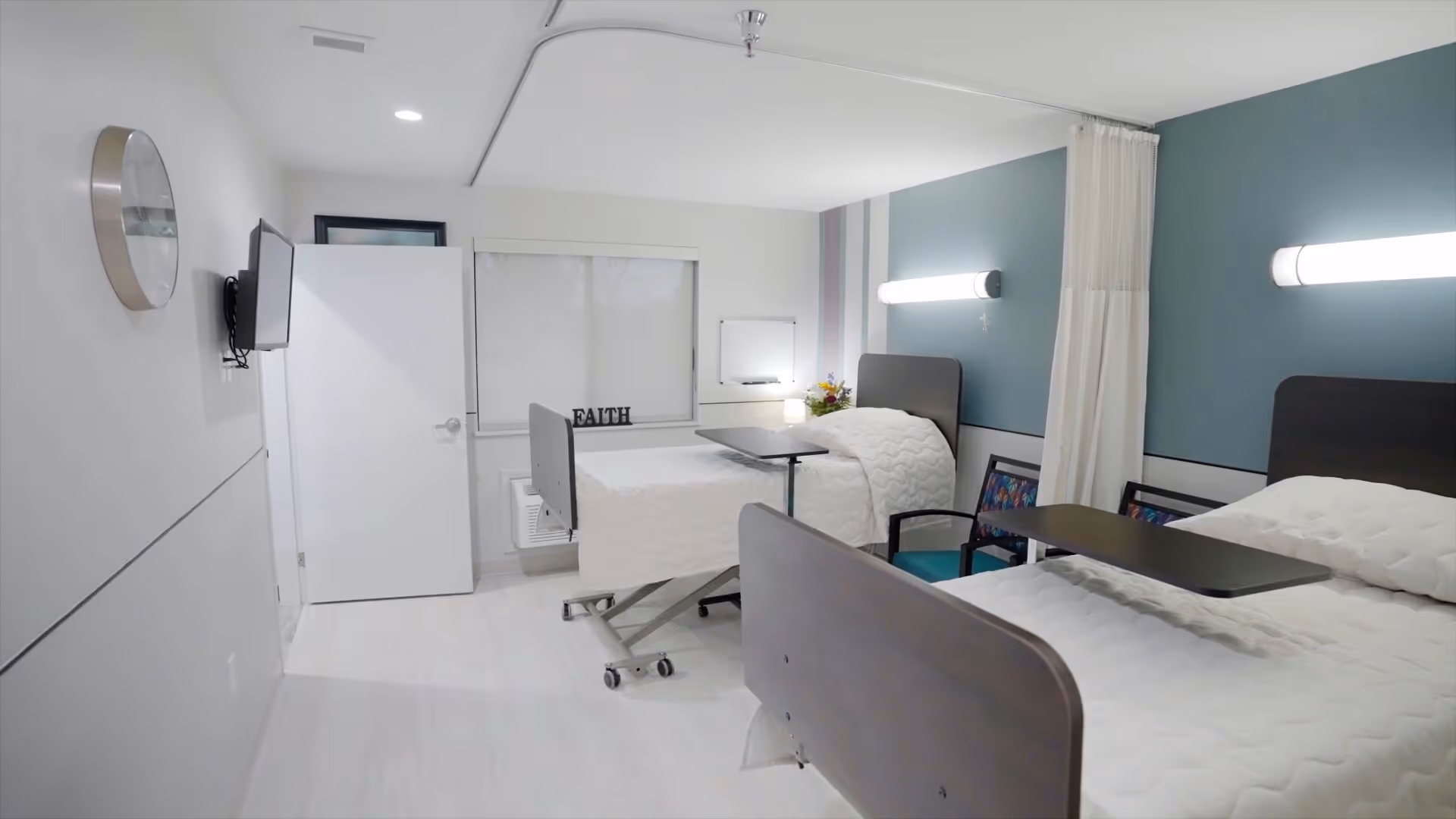 Bright two-bed rehabilitation room with adjustable hospital beds, overbed tables, a curtain divider, wall lights and a wall-mounted TV.