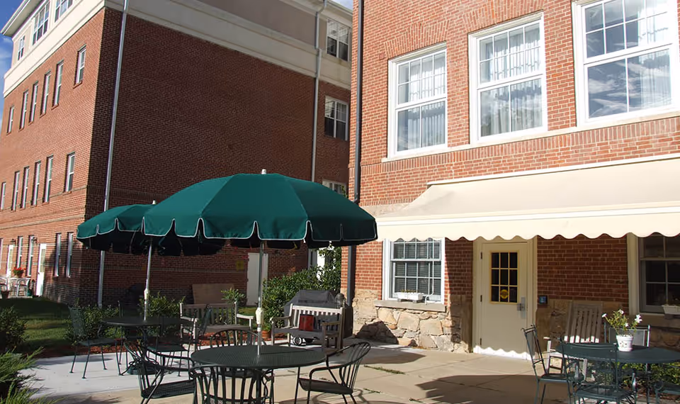 Outdoor patio area at Academy Point at Mystic with green umbrellas shading metal tables and chairs. The patio is adjacent to a brick building with windows and a door under a beige awning.