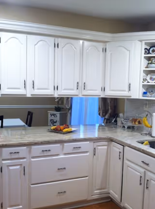 A bright kitchen with white cabinets and drawers, a granite countertop with a fruit plate, and a view into a dining area with a table and chairs. The kitchen has decorative plates on shelves and a window with curtains.