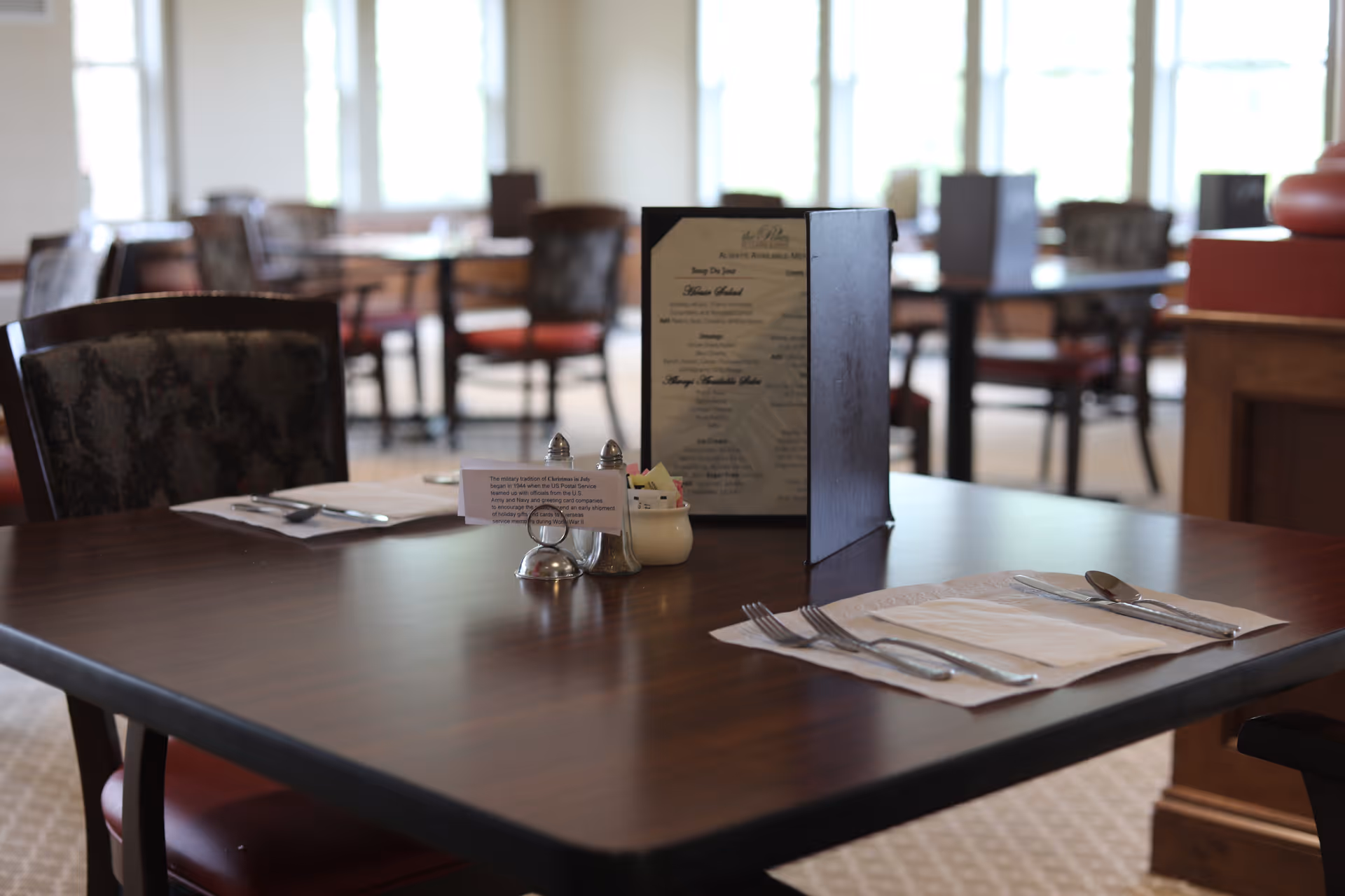Empty dining table set with silverware, napkins, a menu holder and condiments in a senior living dining room.