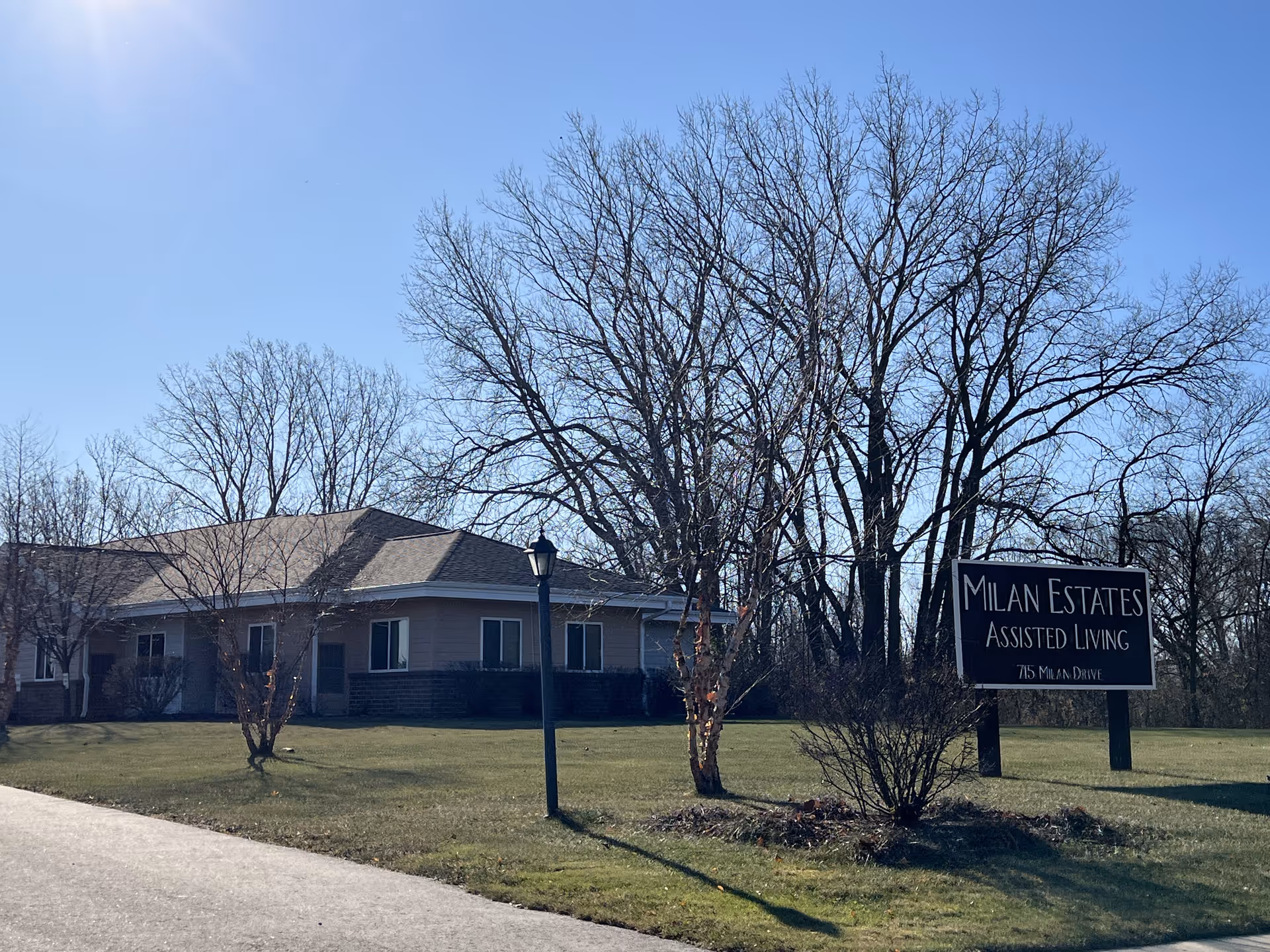 Exterior view of Milan Estates Assisted Living building with a sign in front reading 'Milan Estates Assisted Living 715 Milan Drive'. The building is single-story with a sloped roof, surrounded by leafless trees and a grassy lawn under a clear blue sky.