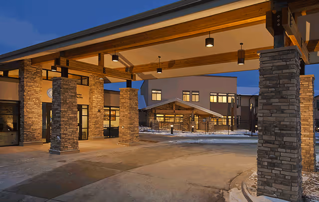 Entrance area of Columbine Commons Assisted Living facility at dusk, featuring stone pillars supporting a wooden roof structure with lights, and the building visible in the background with snow on the ground.