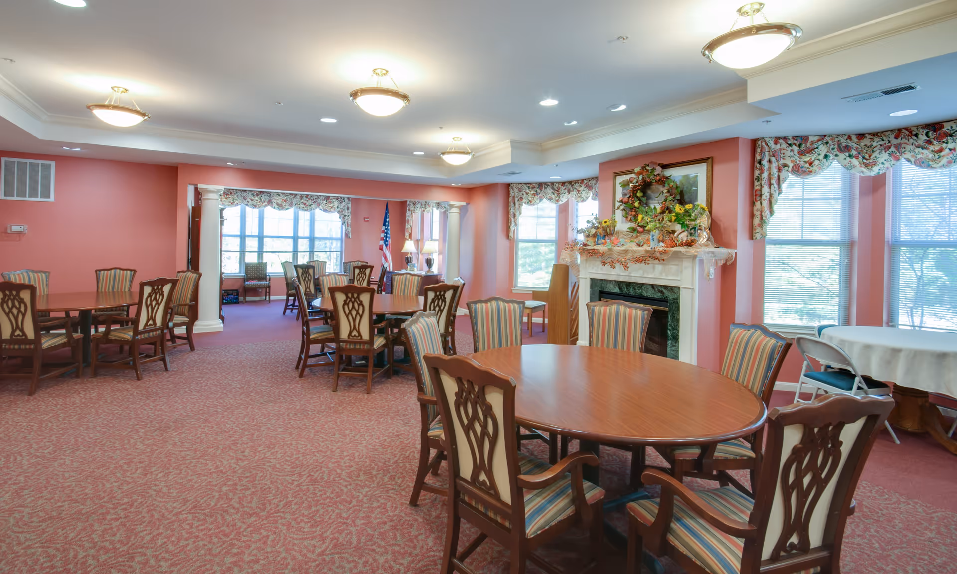 A spacious dining room with multiple wooden tables and chairs with striped upholstery. The room has pink walls, carpeted floors, large windows with floral valances, and a decorated fireplace with a wreath and flowers. Ceiling lights illuminate the room.