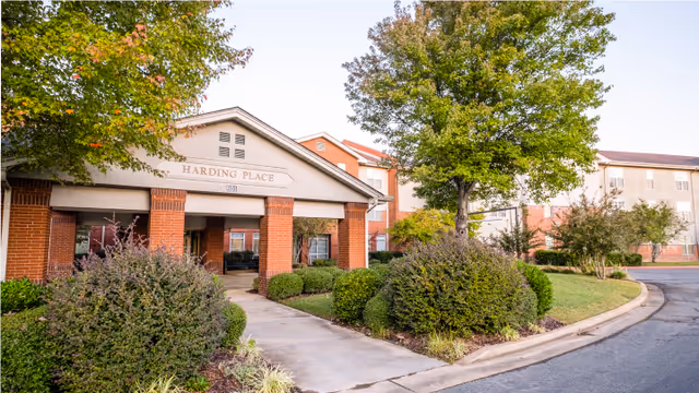 Exterior view of Harding Place Retirement facility showing the entrance with a covered walkway supported by brick columns, surrounded by well-maintained bushes and trees under a clear sky.