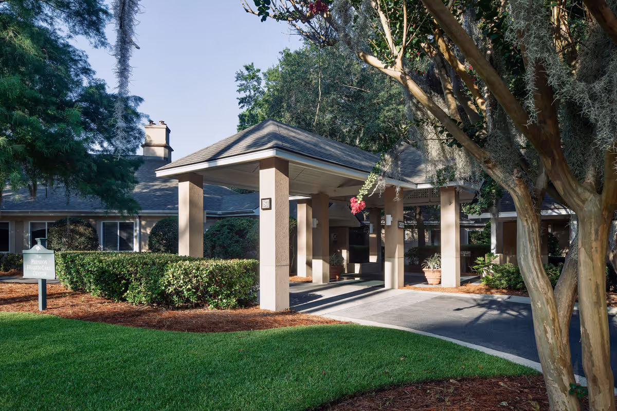 Entrance to a senior living facility with a covered drop-off area supported by beige columns, surrounded by green bushes, trees, and well-maintained grass under a clear sky.