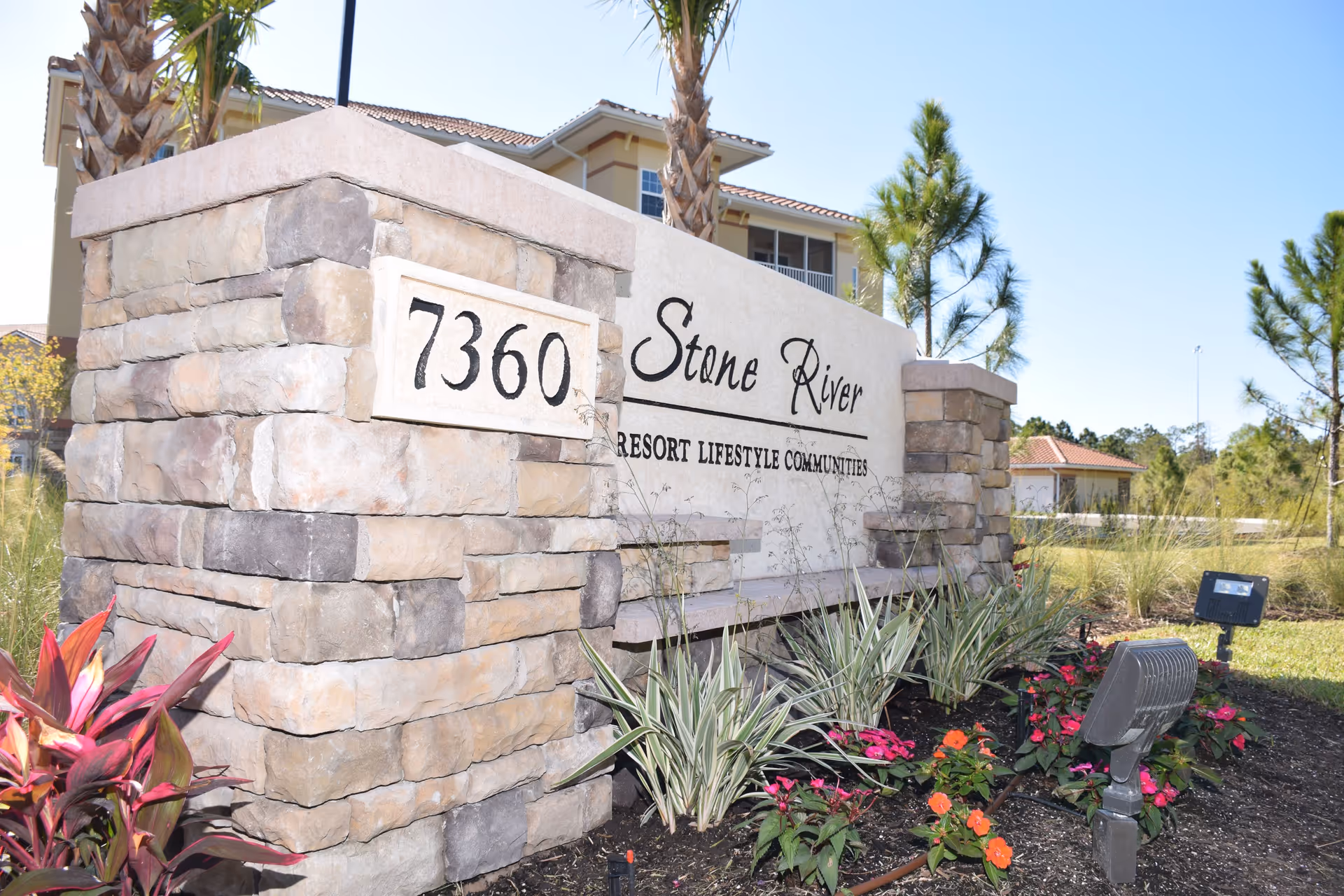 Stone River Retirement Community entrance sign made of stone with the address number 7360 displayed. The sign is surrounded by landscaping with flowers and plants, and a building with a tiled roof is visible in the background under a clear blue sky.