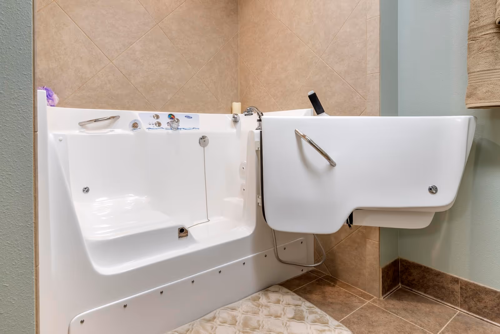 A walk-in bathtub with a side door open, featuring built-in seat, faucet, handrail, and tiled walls in a bathroom setting with a towel hanging on the wall.