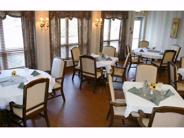 Bright dining room with round tables set with white tablecloths, napkins, glassware and chairs beneath floral curtains.