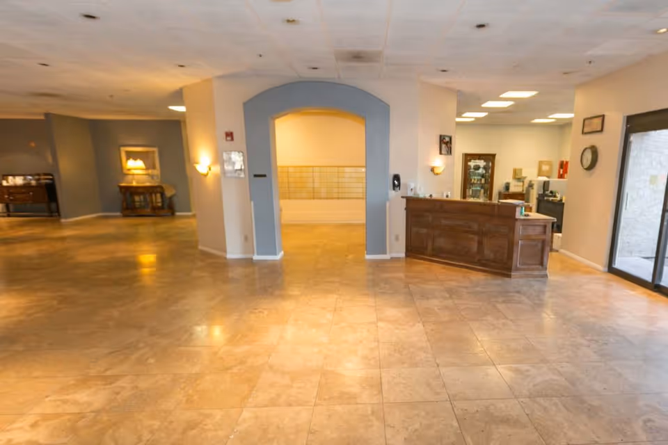 Spacious interior lobby area with tiled floor, a wooden reception desk on the right, an arched doorway in the center, and some furniture and lighting fixtures along the walls.