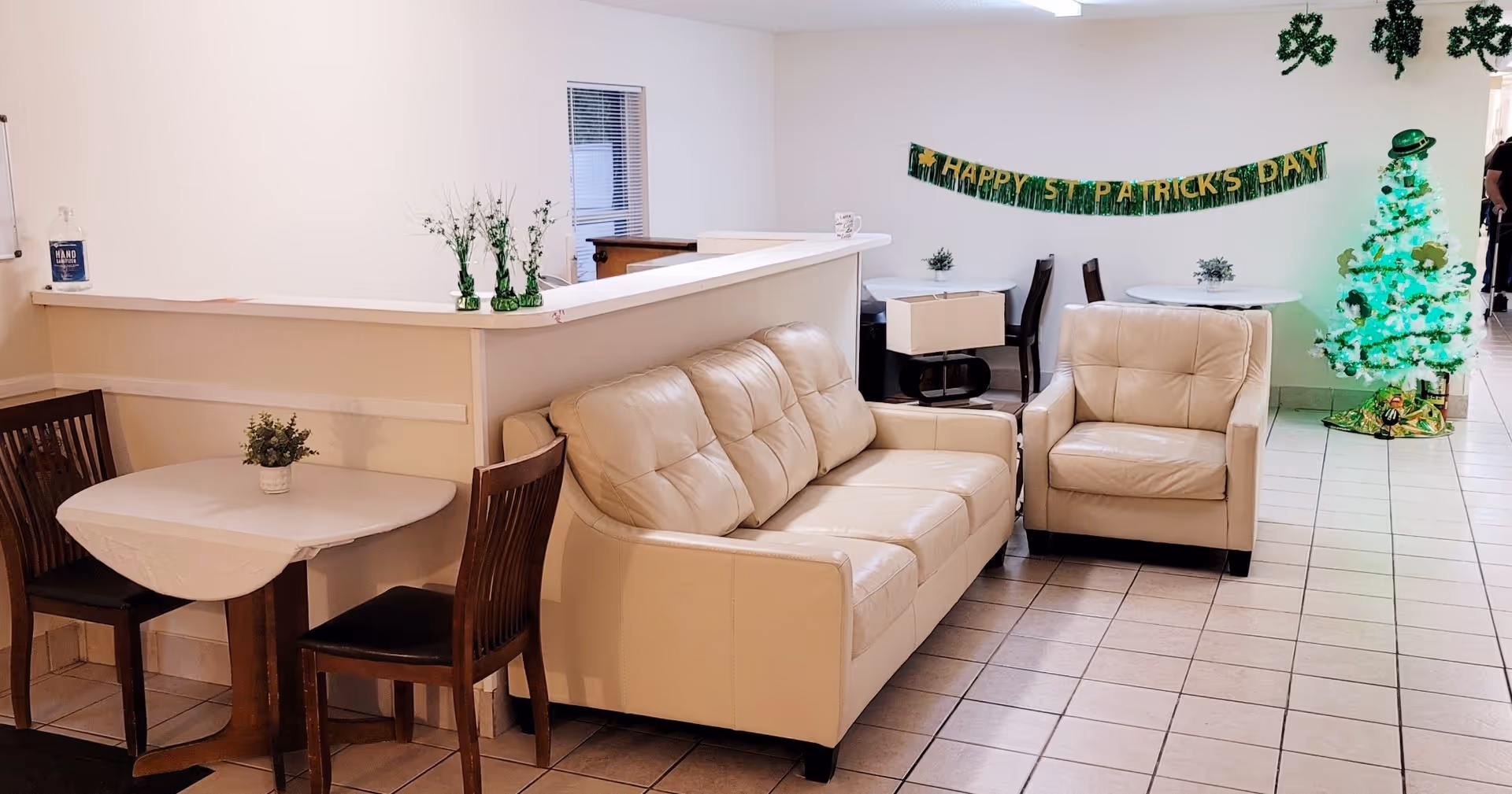 Small communal lounge with a beige leather sofa and armchair, dining tables, and St. Patrick's Day decorations including a green-lit tree.