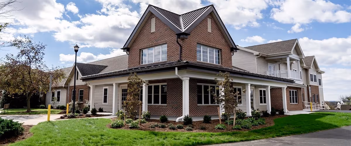Two-story brick-and-siding retirement community building with a covered wraparound porch, landscaped lawn, and a cloudy sky.